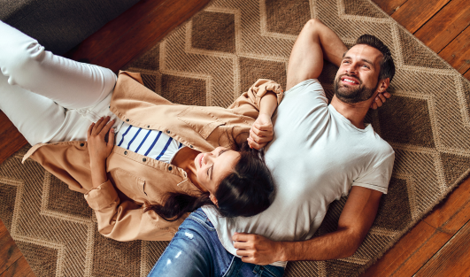 A woman is lying on a man's stomach on a rug as they smile and look up.