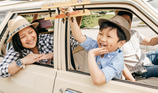 A child sitting in a car smiling while holding an airplane out the window with a woman sitting in the backseat looking at him out the window and smiling.