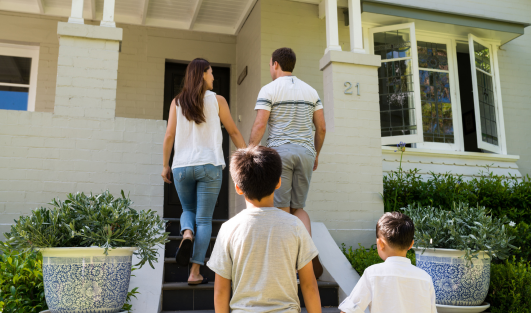 A man and woman holding hands walking up the outside front steps of a home while two kids follow them.