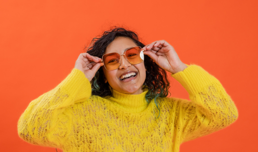 A woman smiling while holding the sides of her sunglasses on her face.