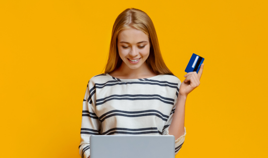 A woman smiling and looking at a laptop while holding a card in one hand.