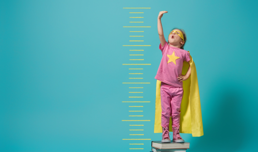 A child standing on a stack of books with a cape and measuring herself as she holds her hand above her head.