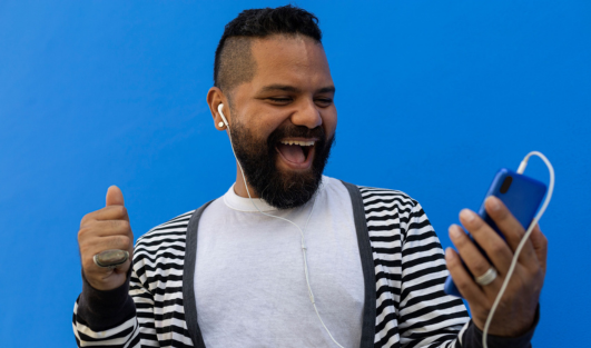 A man with headphones in his ears and a phone in his hand smiling at his device.