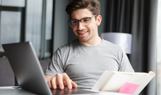 A man sitting down with a notebook smiling at a laptop screen.