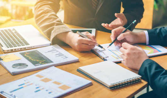 A table filled with charts, a laptop, pens, and paper with two people reviewing a document.