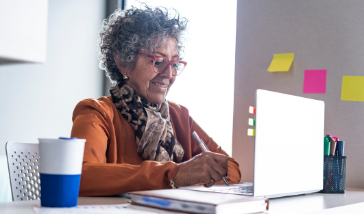 An older woman sitting at a desk with office supplies and a laptop.