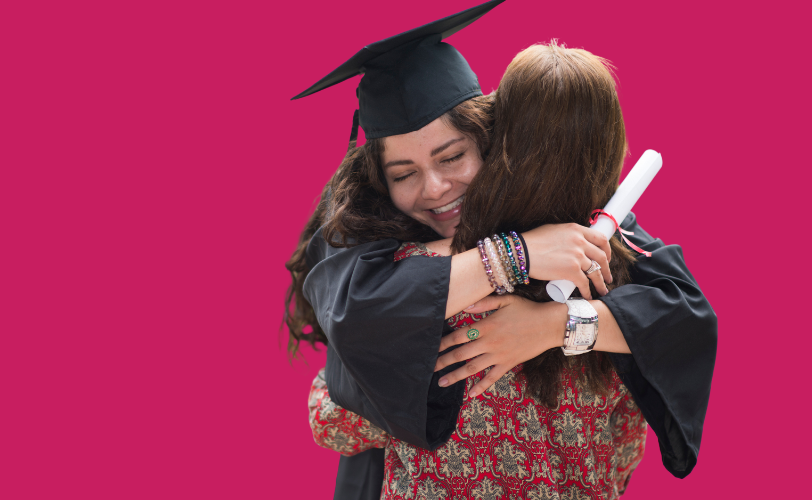 A graduate in a cap and gown hugging a lady while holding a rolled-up diploma.