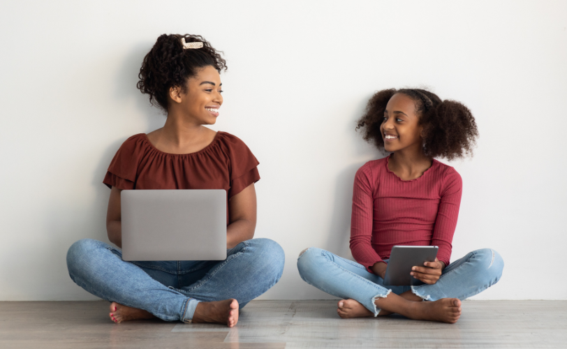 A woman sitting on the floor with a laptop on her lap, looking at a child sitting with a tablet on her lap, smiling back at the woman.