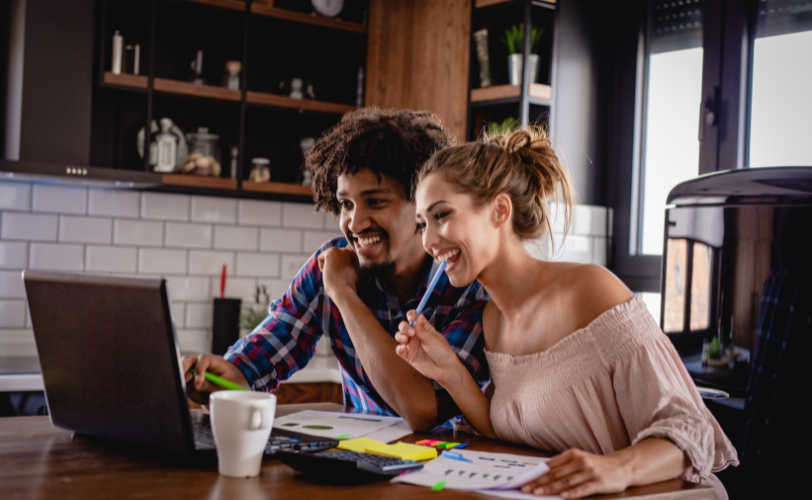 A man and woman in a kitchen are smiling while leaning over the counter, looking at a laptop.