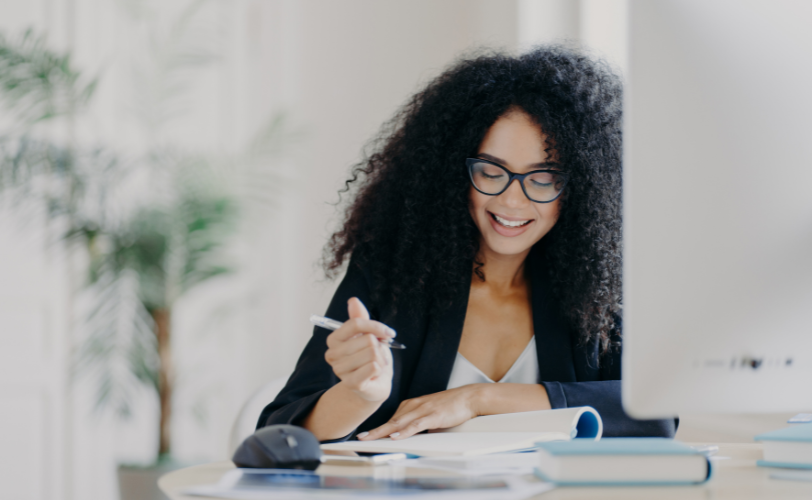 A woman smiling while sitting at a desk holding a pen and looking at her notebook on the desk.