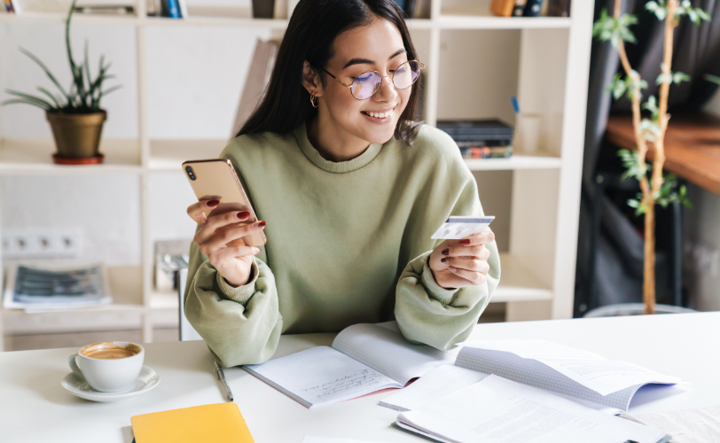 A woman sitting at a table holding and looking at her debit card in one hand while holding her phone in her other hand with notebooks and a mug sitting on the table in front of her.