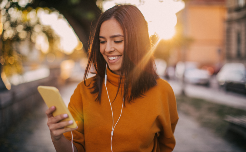 A woman wearing headphones and looking at her phone as she walks outside.