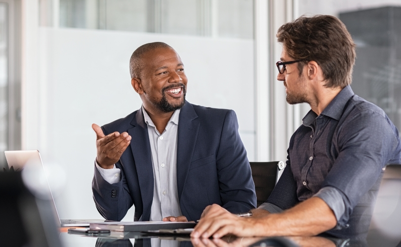Two men are sitting at a table in an office space, smiling and having a conversation.