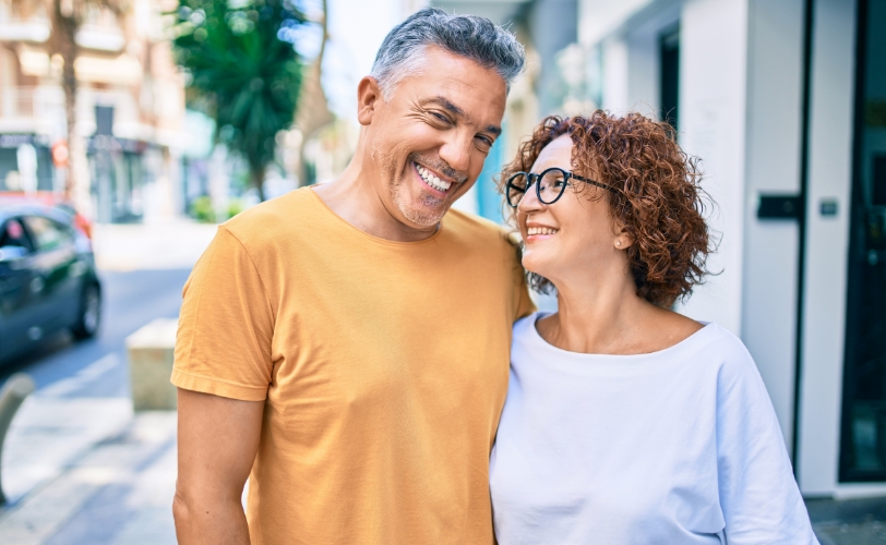 A man and woman smiling with arms around each other as they walk down the street outside.