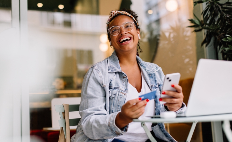 A woman smiling while sitting at a table with a laptop on it, with wireless headphones in her ears, holding a phone in one hand and a credit card in another.