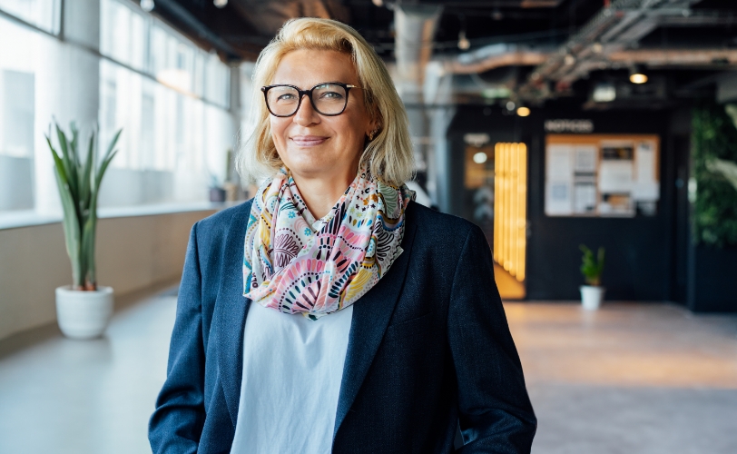 A woman smiling wearing a decorative scarf and glasses in an industrial building with windows.