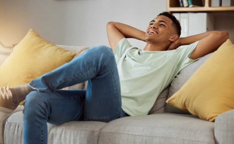 A man smiling and relaxing on a couch with his hands behind his head and legs crossed.