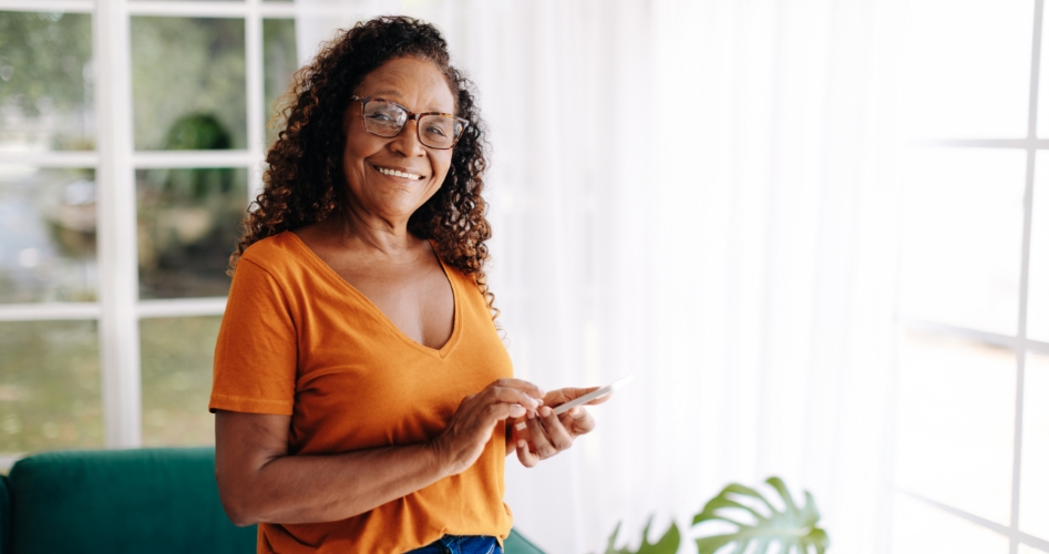 A woman is smiling and holding a phone in her hand next to big windows.