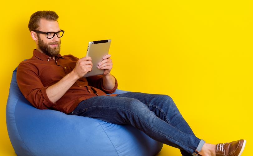 A man smiling and sitting on a bean bag chair holding a tablet and looking at it.