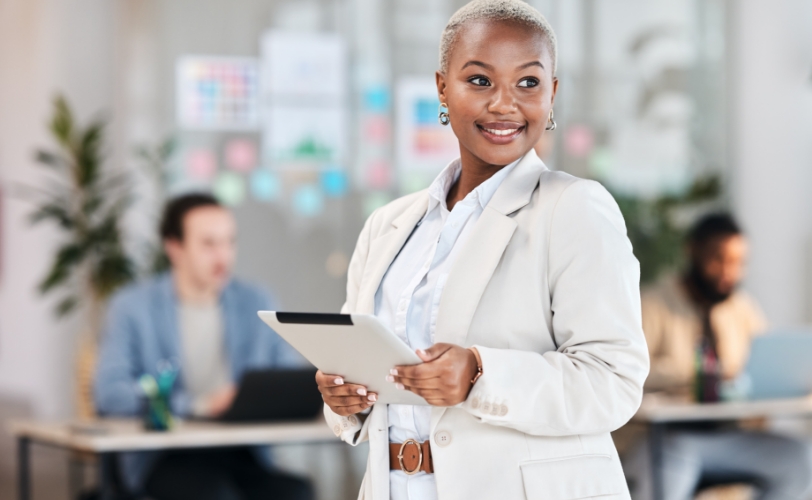 A woman standing in a professional setting while holding a tablet and looking off in the distance.