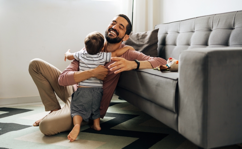 A man sitting on the floor next to a couch, smiling while hugging his child.