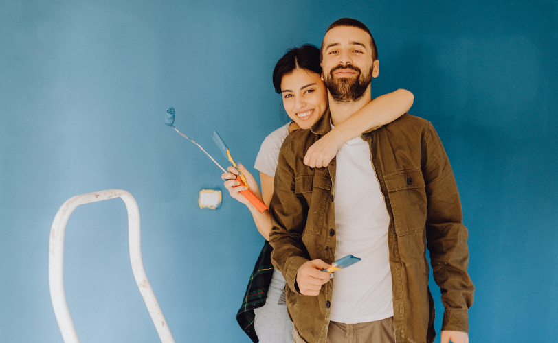 A woman hugging a man from behind holding a paint roller and paint brush.