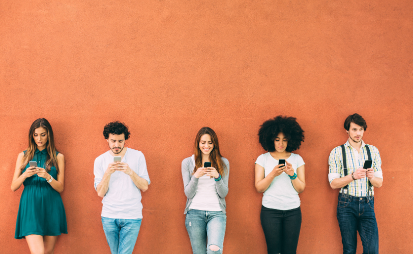 A line of people standing against a wall and typing on their phones.