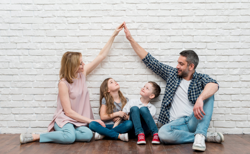 A man and woman sitting on each side of two kids and making a house with their hands around them.