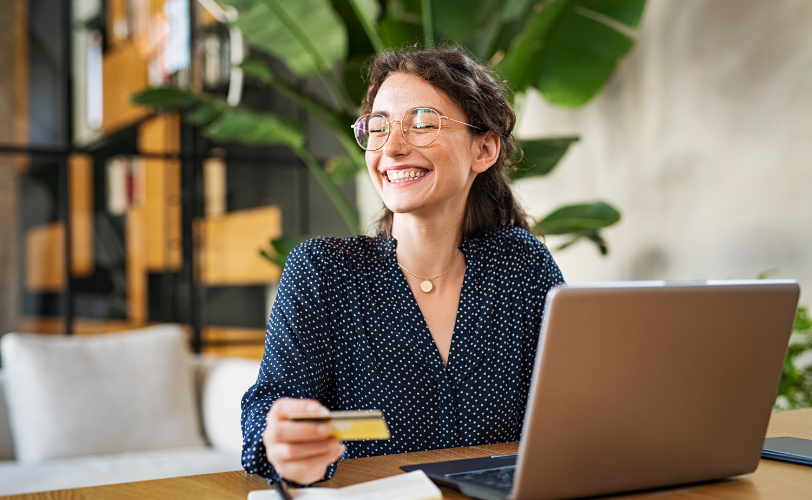 A woman sitting at a table is smiling and looking off in the distance while she holds a credit card and sits a laptop in front of her.