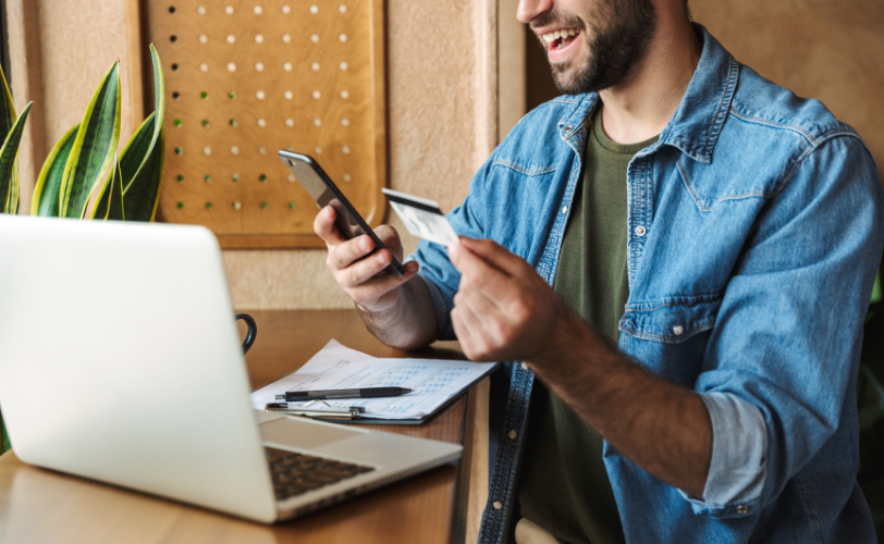 A man is sitting at a table, looking at his phone and credit card in his hands. His laptop, pen, and paper are also on the table in front of him.