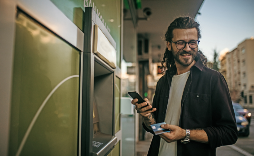 A man holding his card and phone standing next to an ATM outside in a city.