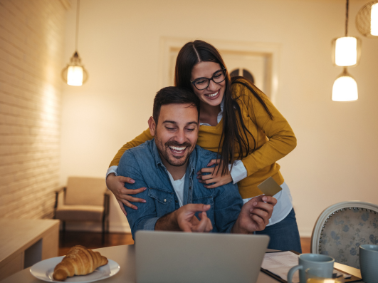 A man and woman happily looking at a laptop together at a table with cups and food next to them.