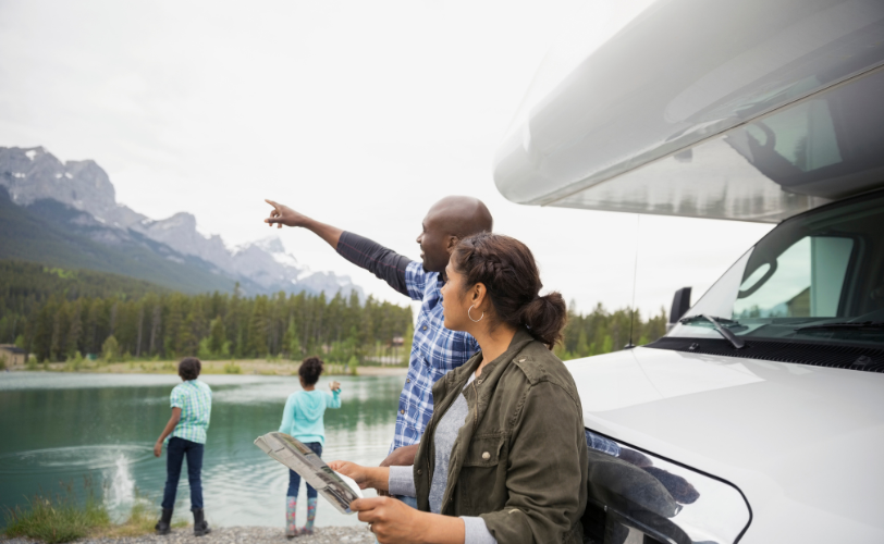 A man and woman looking towards the mountains in front of an RV with two kids nearby skipping rocks in the water.