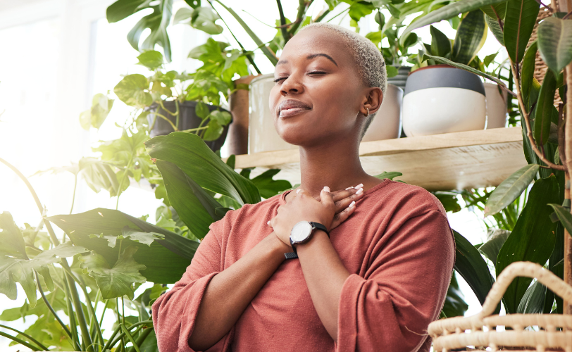 A woman in a room full of plants is crossing her hands on her chest with her eyes closed.