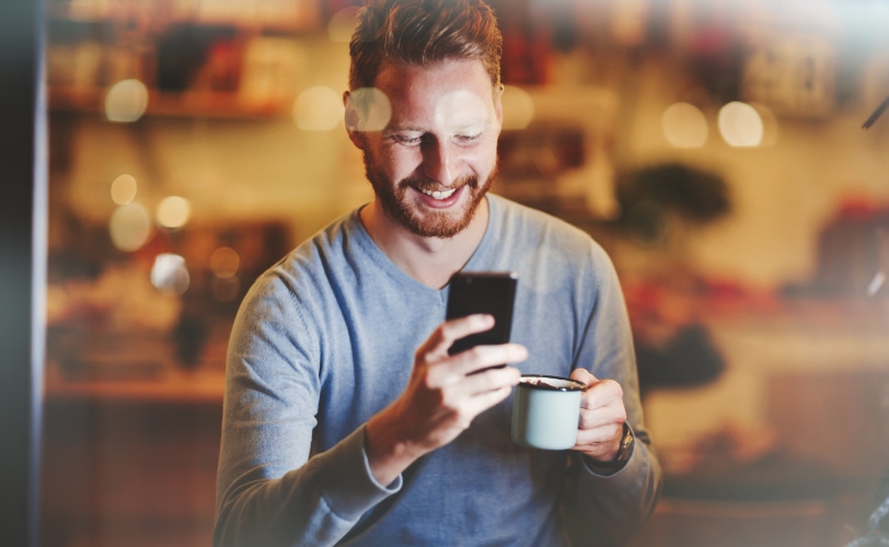 A man is smiling, holding a mug, and holding his phone while looking at it in a coffee shop.