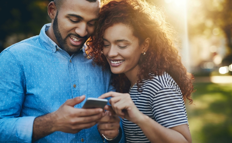 A man and woman smiling and looking at a phone together outside.