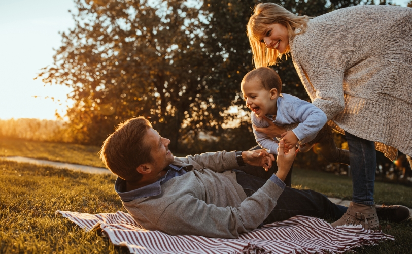 A man happily lying outside on a blanket, sitting up to hold a child while another woman happily passes a child to the man.