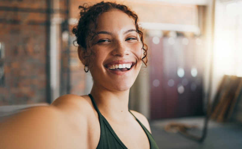 A woman taking a selfie and smiling in a building with the sun shining behind her.