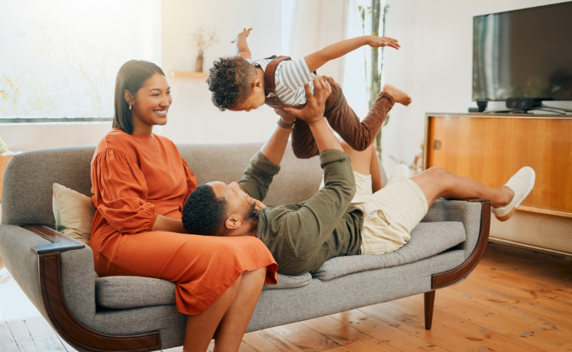 A man, woman, and child on a couch, with the man flying the child like an airplane.