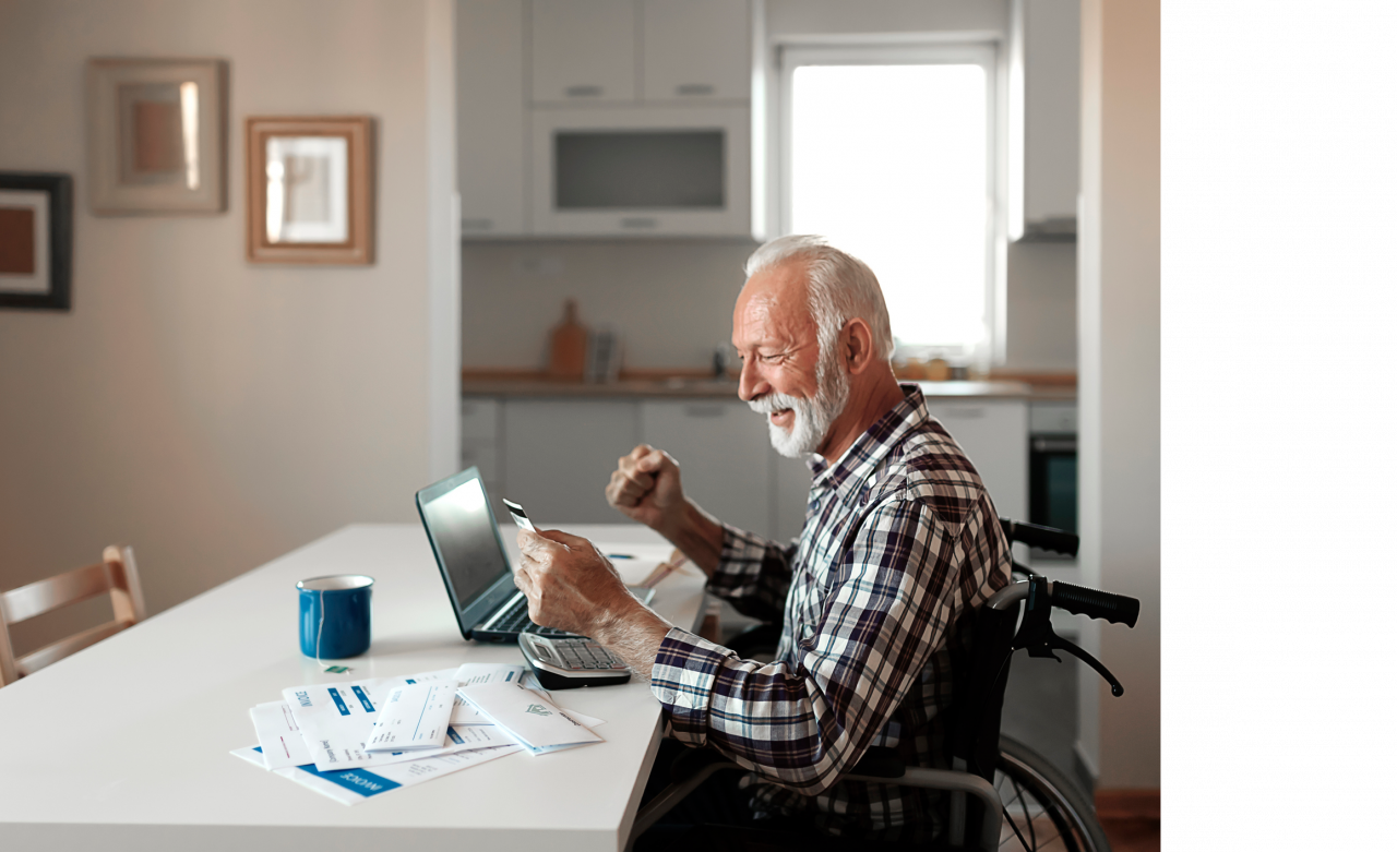 An older man in a wheelchair sitting at his dining room table at home on his laptop with a credit card. 