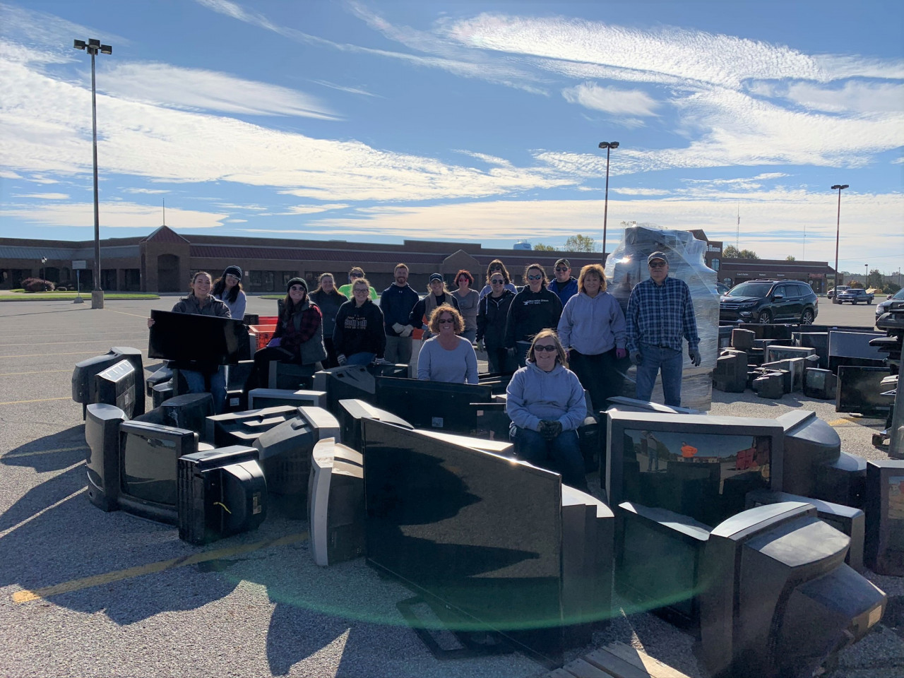 A big group of people in a parking lot with a bunch of TVs and electronics collected around them for electronic recycling day.