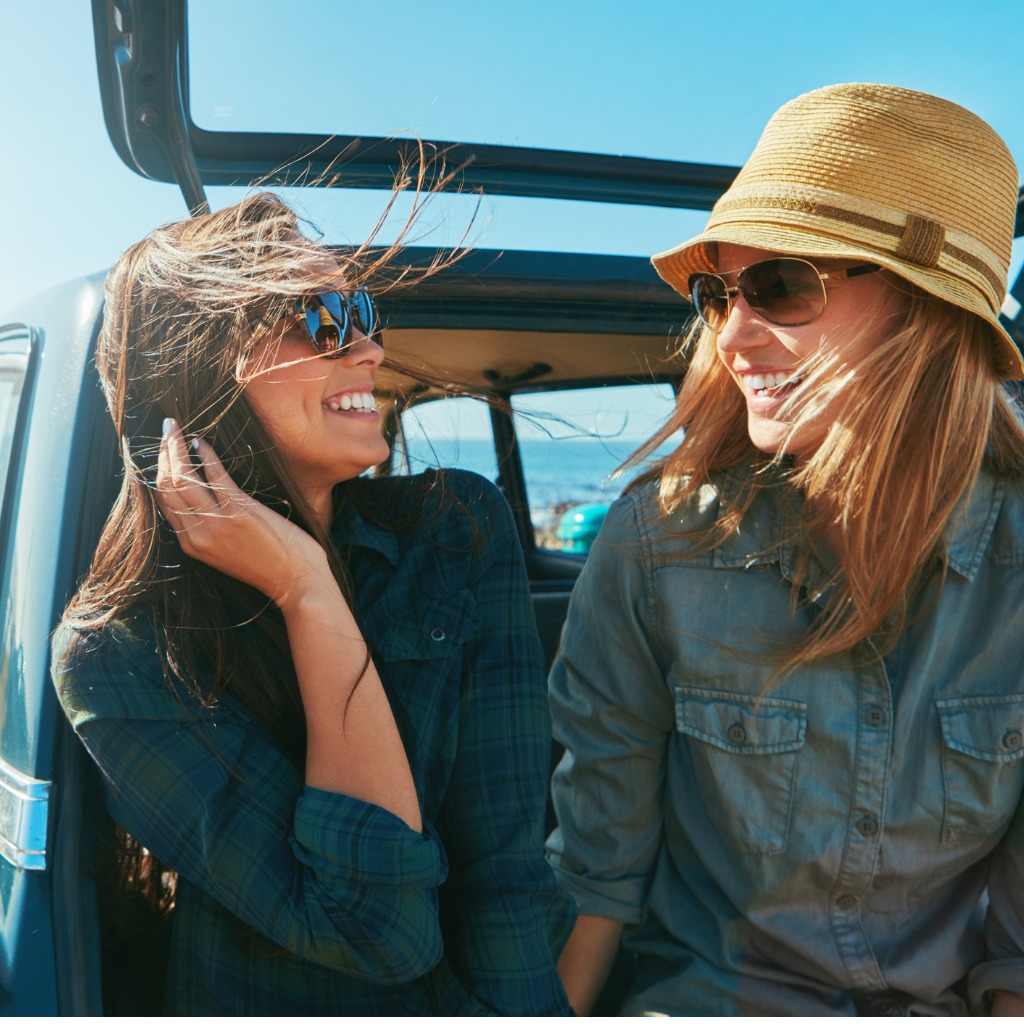 Two women sitting in the trunk of a car on a beach with their hair blowing in the wind and enjoying each other's company.