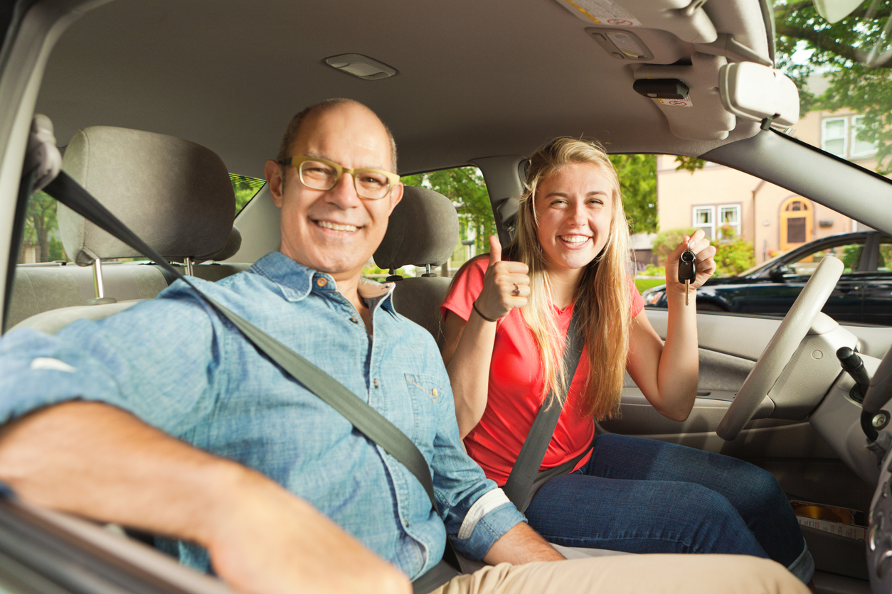 A woman in the driver's seat of a vehicle holding keys and giving a thumbs up because she got her first car and her dad is in the passenger's seat smiling.