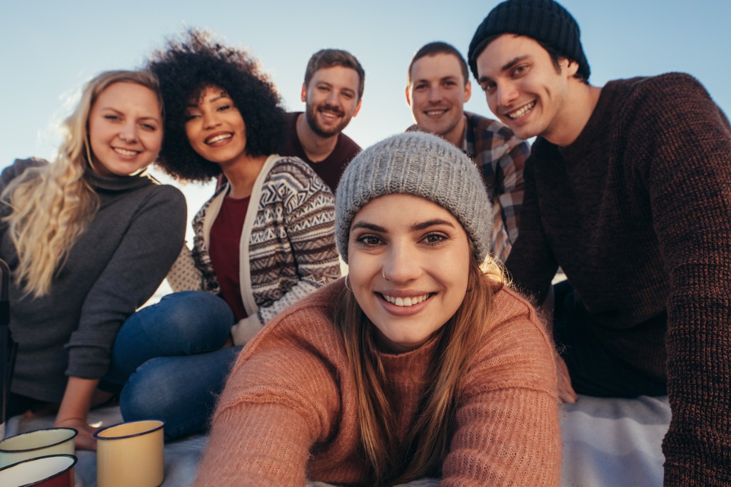 Woman taking a selfie with a group of friends in a sweater and hat on the beach.