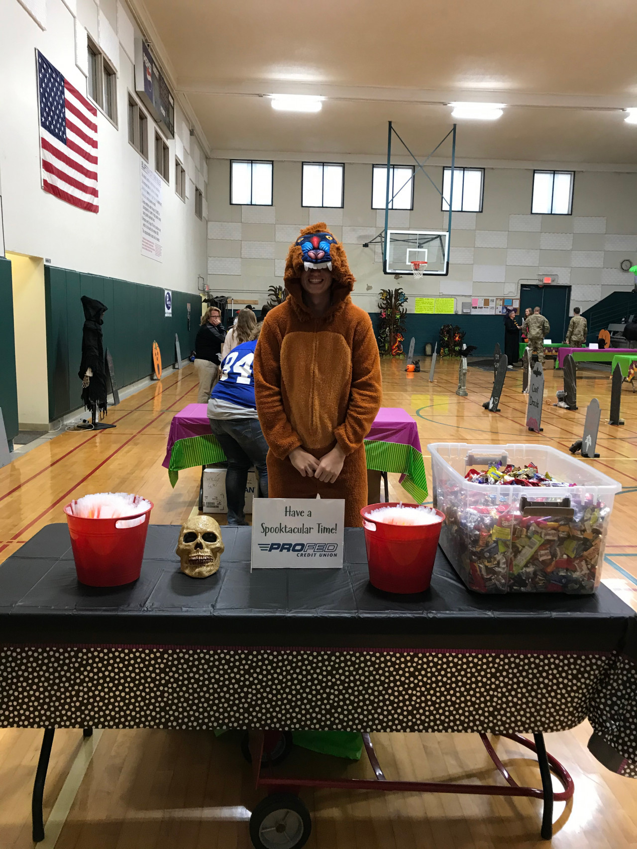 A boy in a chimpanzee costume standing behind a table passing out candy for Halloween in a gymnasium.