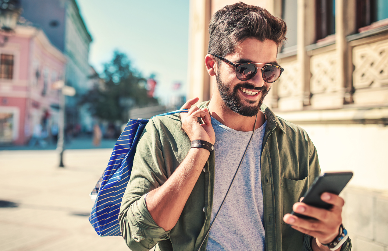 A young man enjoying shopping in town with sunglasses on while on his mobile device.