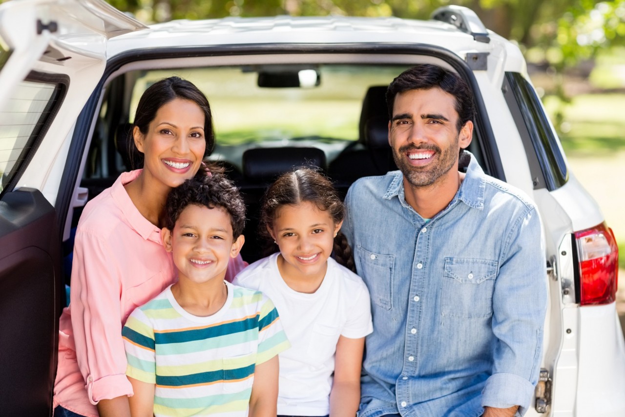 A family smiling while sitting in the trunk of their vehicle on a sunny day.