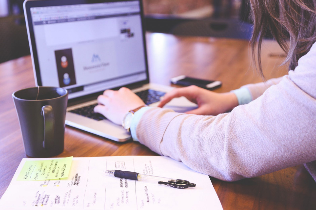 Someone sitting a desk typing on a laptop with a coffee mug and papers sitting next to them.