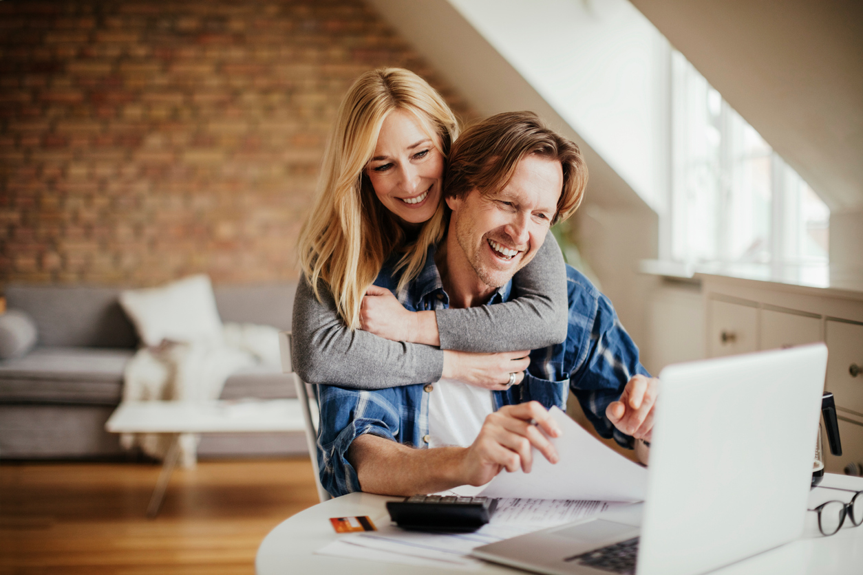 A woman hugging a man from behind while he is sitting down and they are both smiling while looking at a laptop full of future financial plans.