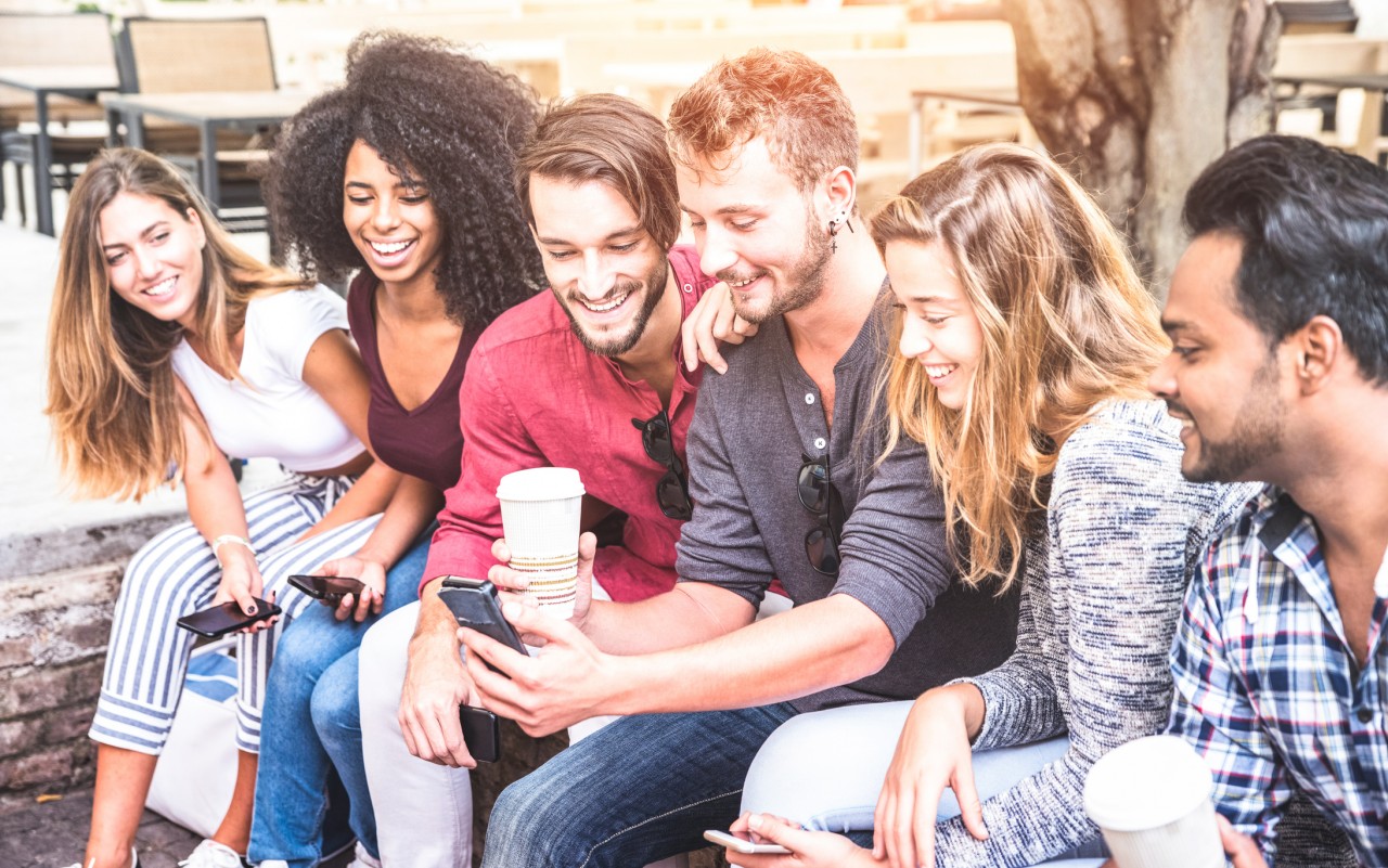 A group of friends sitting outside under a tree while huddled around a phone, drinking coffee, and Facetiming friends together.